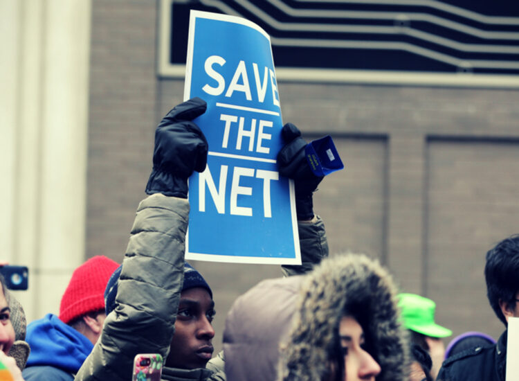 A person at a protest holds a sign reading "Save the Net"