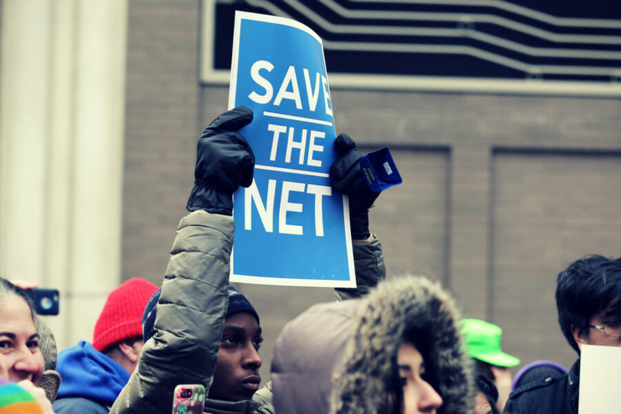 A person at a protest holds a sign reading "Save the Net"