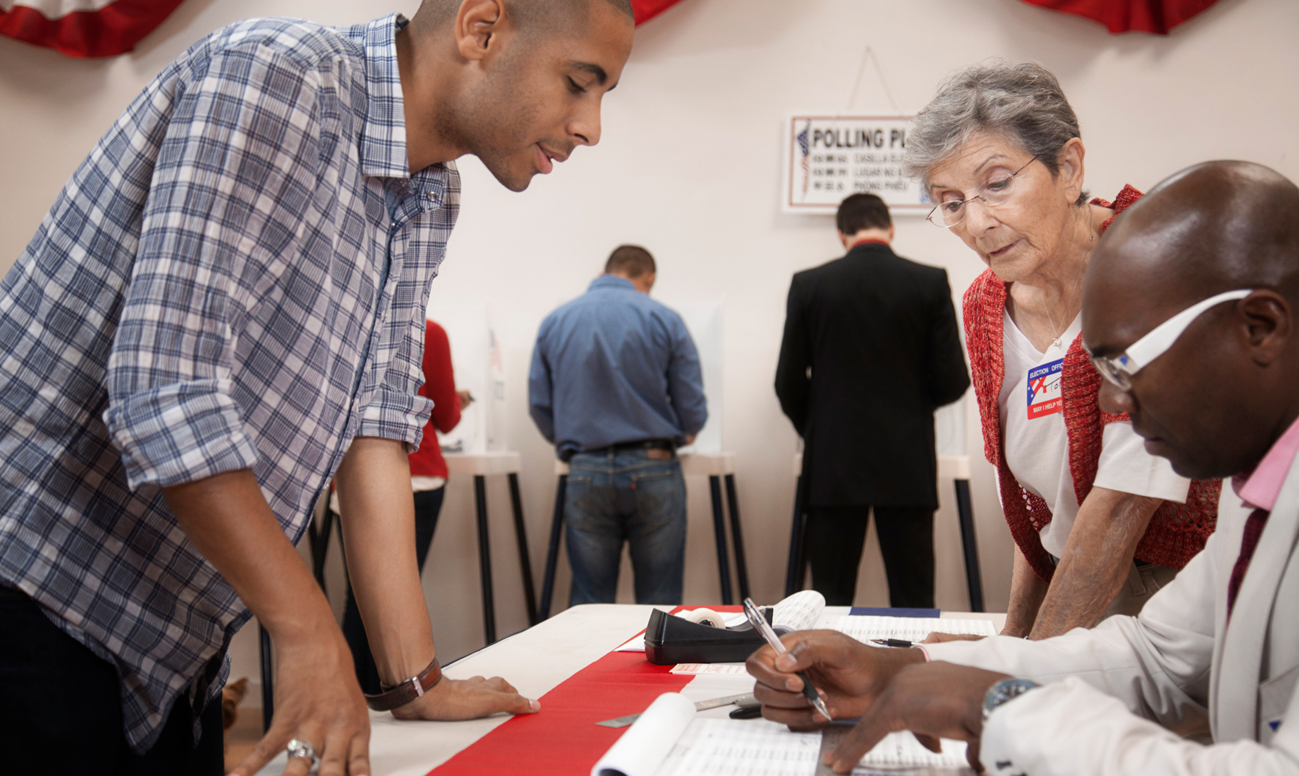 Pollworkers check a man's voter registration status while other people fill out their ballots in the background.