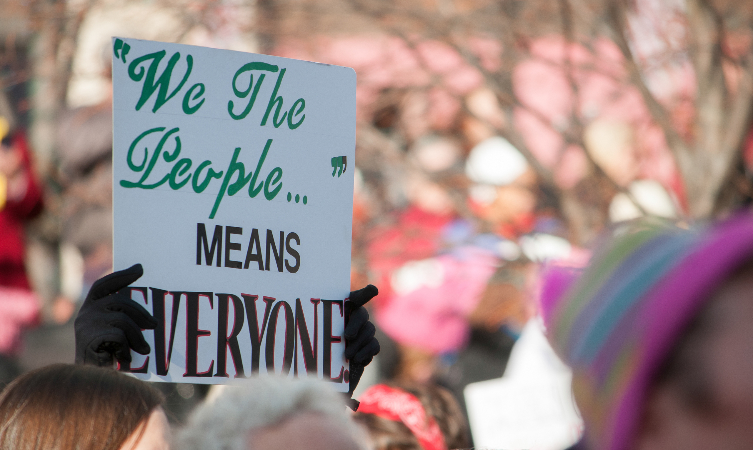 A sign held among a crowd says "'We The People' means everyone"